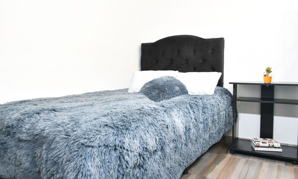 Un lit recouvert d’une couverture bleue moelleuse, avec une tête de lit noire en tissu, des oreillers blancs et une petite table sur laquelle se trouvent une plante et des livres.