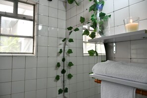 Une salle de bain avec des murs recouverts de carreaux blancs, une fenêtre, une étagère avec une bougie et une plante.