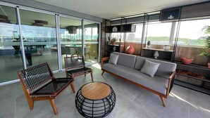 A modern living room with a sofa, two chairs, a coffee table, and a large window with a view of the city. - Studio com vista Mar em Maceió - Max Rev (Macei ó)