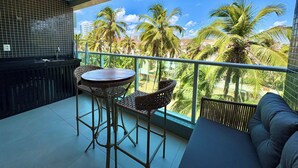 A balcony with a view of palm trees and buildings, a round table with chairs, and a sofa. - Studio com vista Mar em Maceió - Max Rev (Maceió)