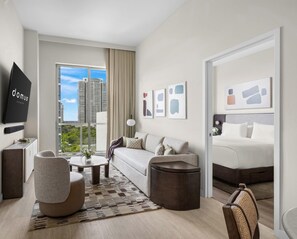 A modern hotel room with a sofa, armchair, coffee table, and a bed. There is a large window with a city view, and artwork on the walls. - Relaxing 1BR Condo at Domus Brickell (Miami)