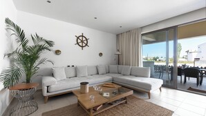 Un salon moderne avec un canapé d’angle, une table basse en verre, une plante en pot et une vue sur l’extérieur.