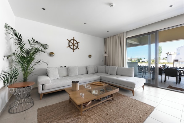 Un salon moderne avec un canapé d’angle, une table basse en verre, une plante en pot et une vue sur l’extérieur.