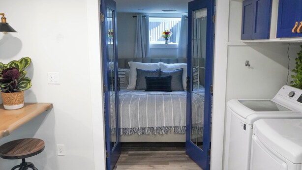 Un couloir étroit mène à une chambre à coucher qui contient un lit, une fenêtre avec des rideaux, un lave-linge et une plante posée sur une étagère.