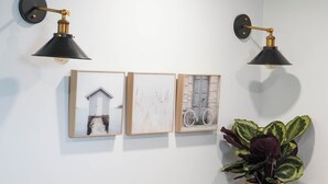 Une pièce comprenant un banc en bois, deux tabourets, des lampes fixées au mur, des œuvres d’art encadrées et une plante en pot.