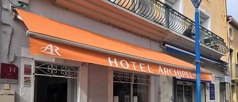 A hotel named 'HOTEL ARCHIPEL' with a prominent orange awning, a visible entrance, and a balcony with a metal railing.