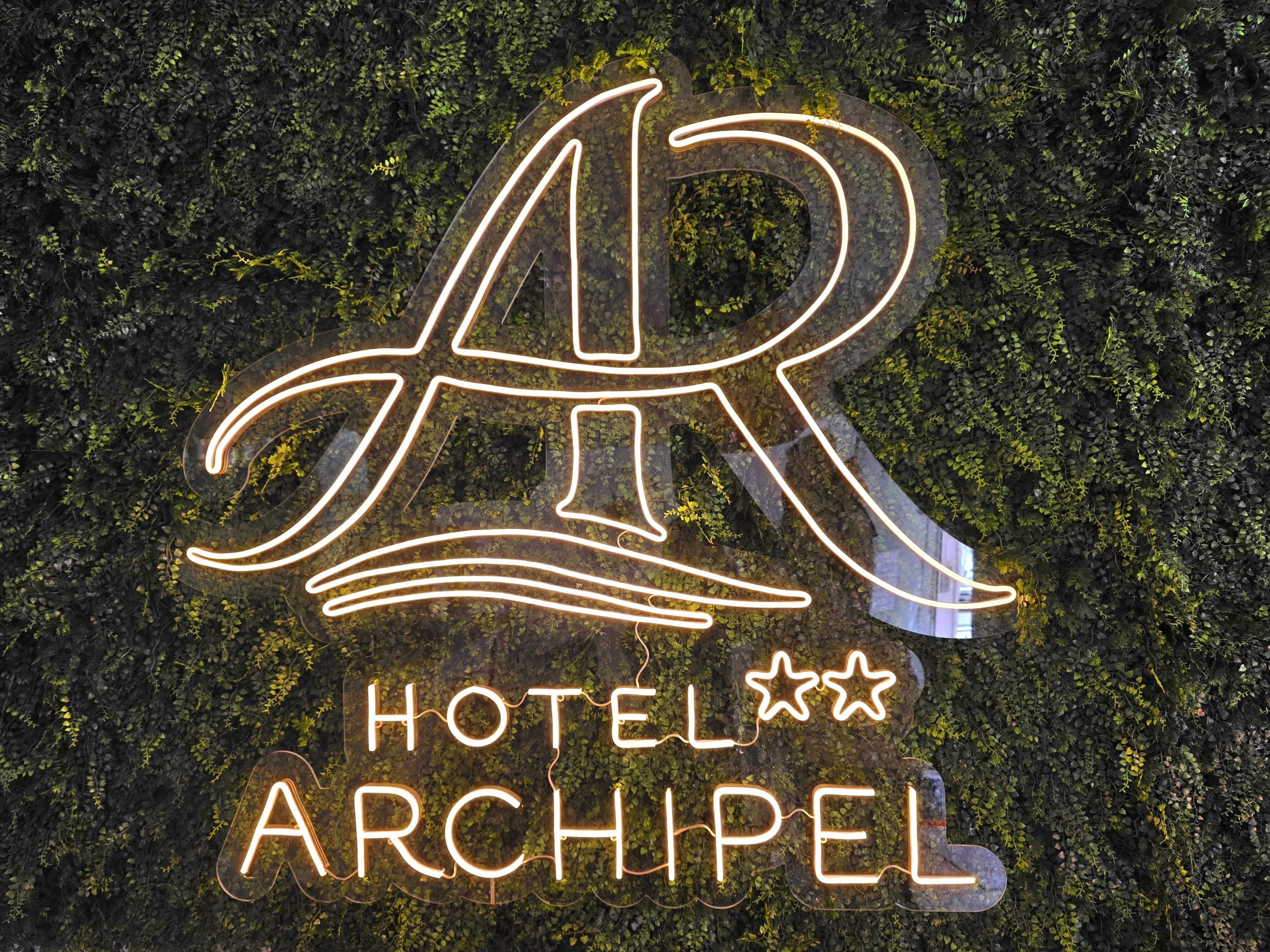 A neon sign with the letters 'AR' and the words 'HOTEL ARCHIPEL' on a wall covered with greenery.