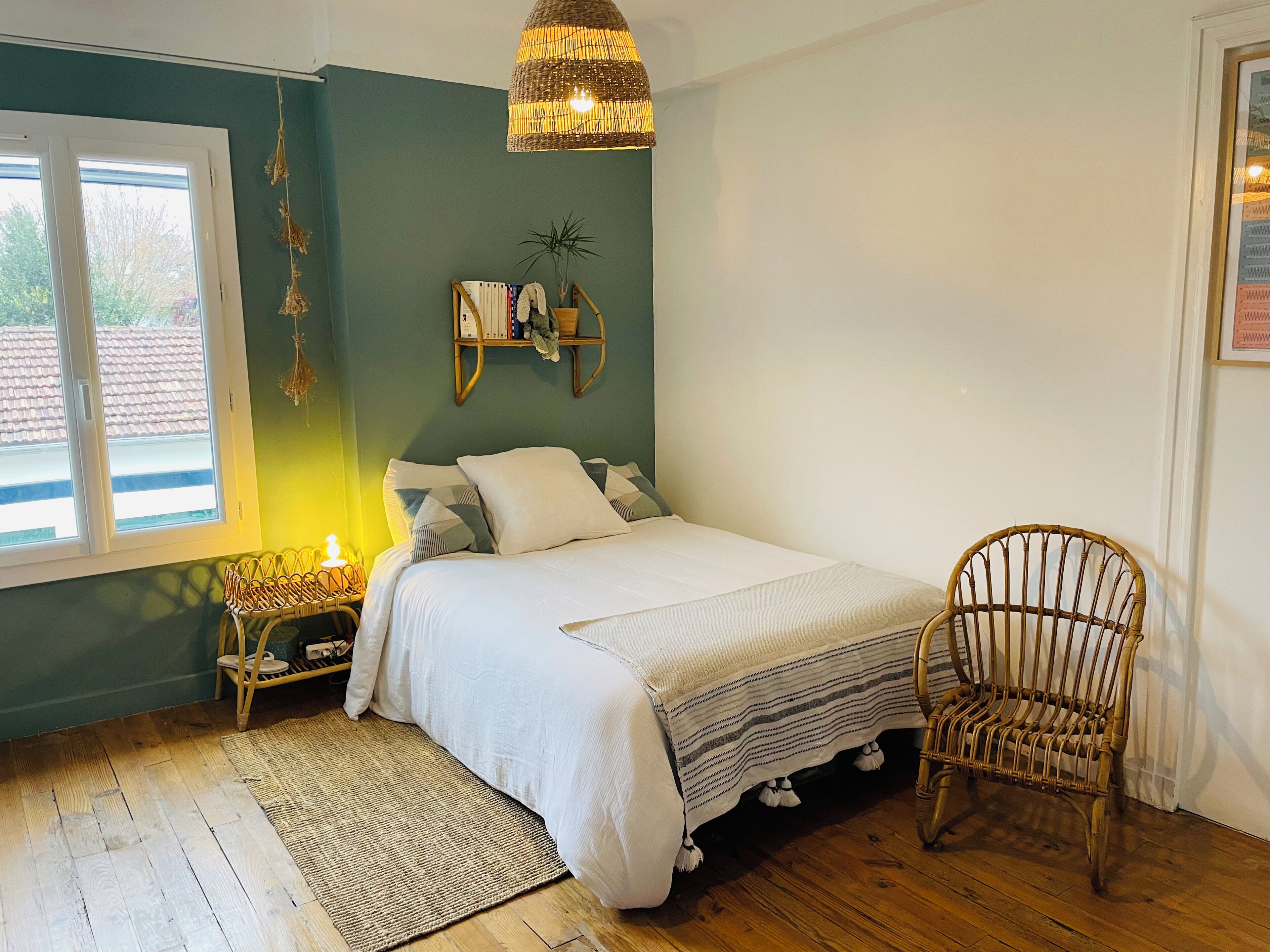 A bedroom with a bed, a wicker chair, a bedside table with a lamp, a hanging plant, and a window with blinds.