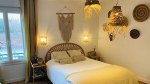 A bedroom with a bed, bedside lamps, a macrame wall hanging, and a wicker chair.
