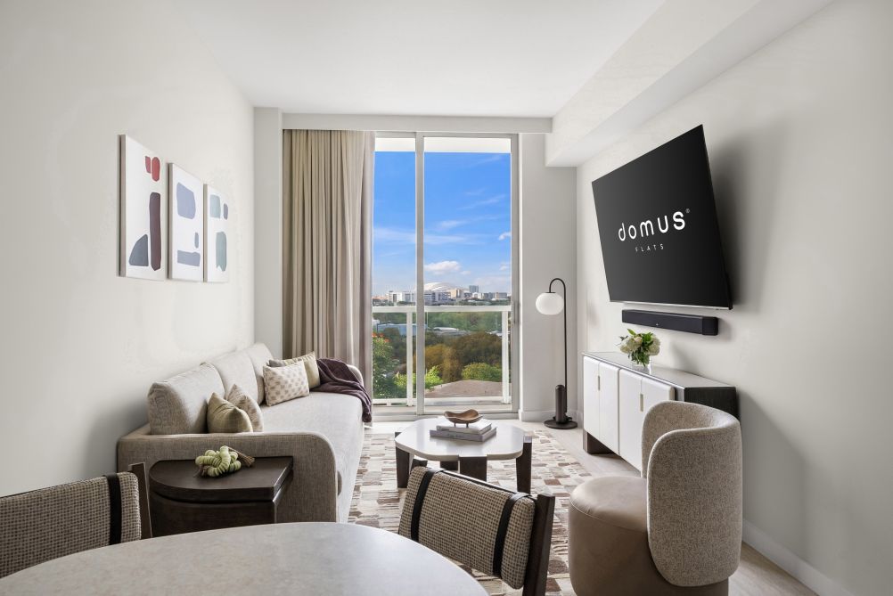 Ein modernes Hotelzimmer mit einer Sitzecke, einem Esstisch, einem Flachbildfernseher und Blick auf die Stadt durch ein großes Fenster.