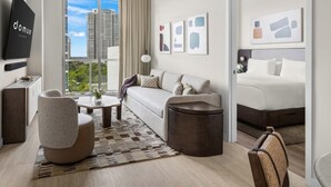 A modern hotel room with a sofa, armchair, coffee table, and a bed. There is a large window with a city view, and artwork on the walls. - Domus Brickell Retreat 1BR Condo (Miami)