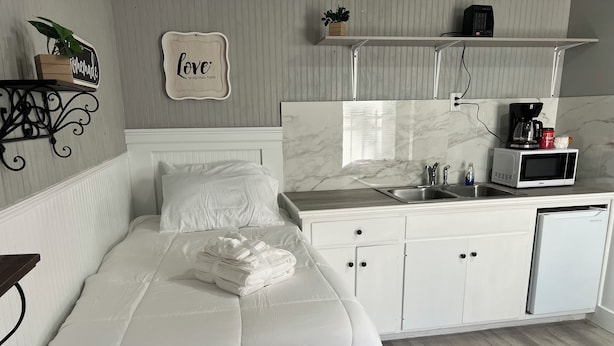A bedroom with a bed, a bedside table, a nightstand, a wall-mounted shelf with a plant, a sign, and a kitchen area with a microwave, coffee maker, and sink.