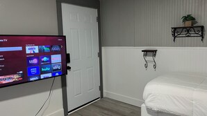 A room with a TV displaying a streaming service interface, a white door, a bed with white bedding, a small shelf with a plant, and a wall-mounted shelf.