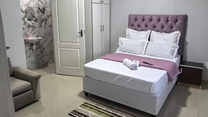 Ein Schlafzimmer mit Bett, Kleiderschrank, Nachttisch, Sessel und einem Badezimmer mit Waschbecken. An der Wand hängt ein Gemälde.