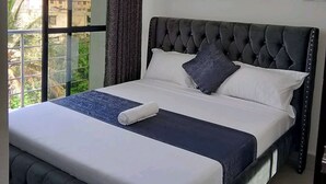 A bedroom with a large window, a bed with a tufted headboard, and a view of buildings outside. - White Palms Apartment Bamburi (Mombasa)