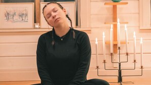 A person sitting cross-legged on a mat in a room with wooden floors, a window, and a menorah with candles. - Lapland Nature & Health (Junosuando)
