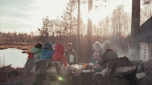 Eine Gruppe von Menschen sitzt bei einem Lagerfeuer in einem bewaldeten Gebiet, im Hintergrund ist eine Holzhütte zu sehen.