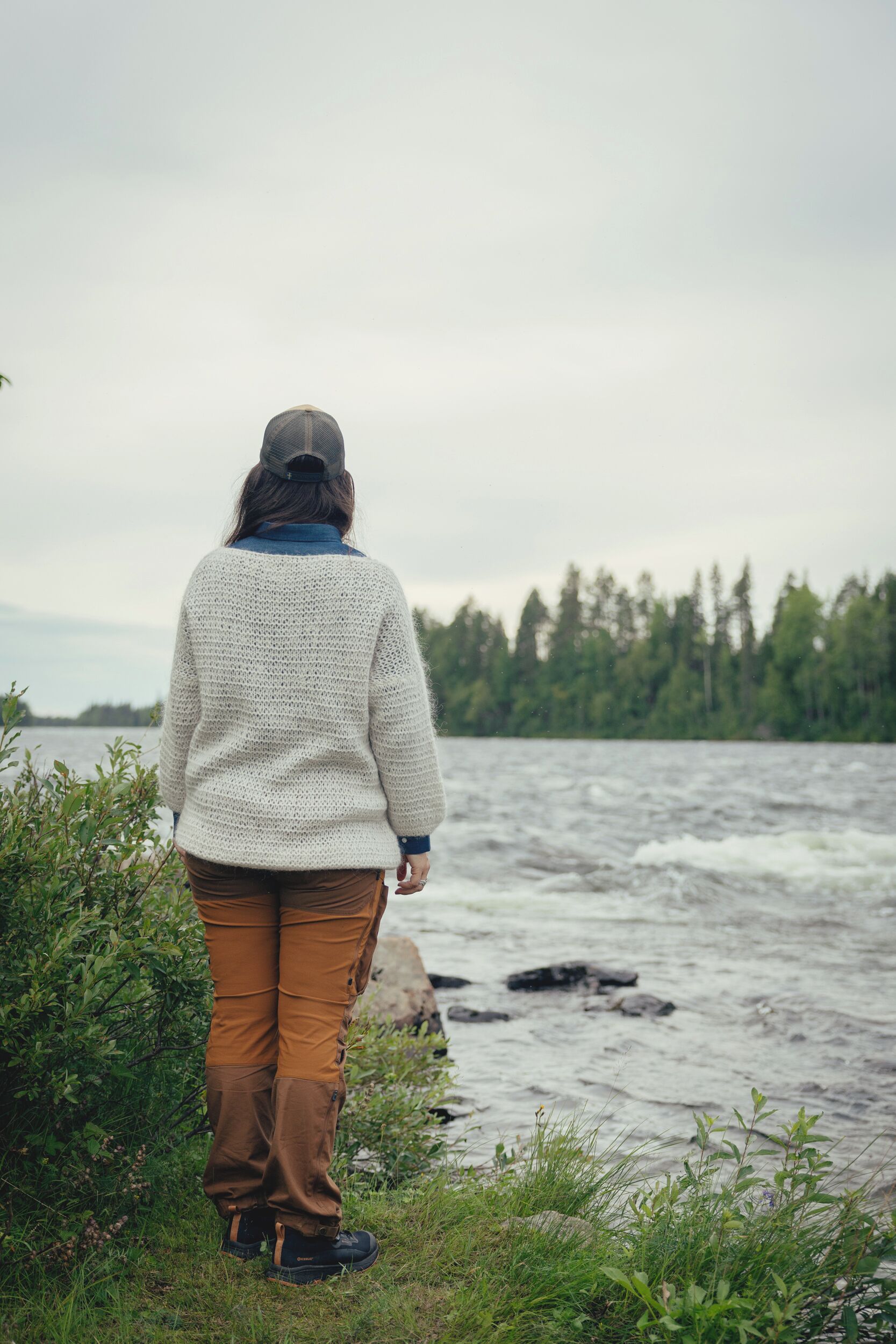 A person standing by a river, wearing a white sweater, brown pants, and a cap.