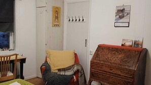 Ein Zimmer mit Holzbalkendecke, einem Bett mit grün gemustertem Bettbezug, einem Holzschrank, einem Sessel mit Decke, einem kleinen Tisch mit Stuhl und einer Tür mit Haken.