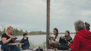 A group of people sitting around a campfire by a lake, with a view of trees in the background. - Lapland Nature & Health (Junosuando)