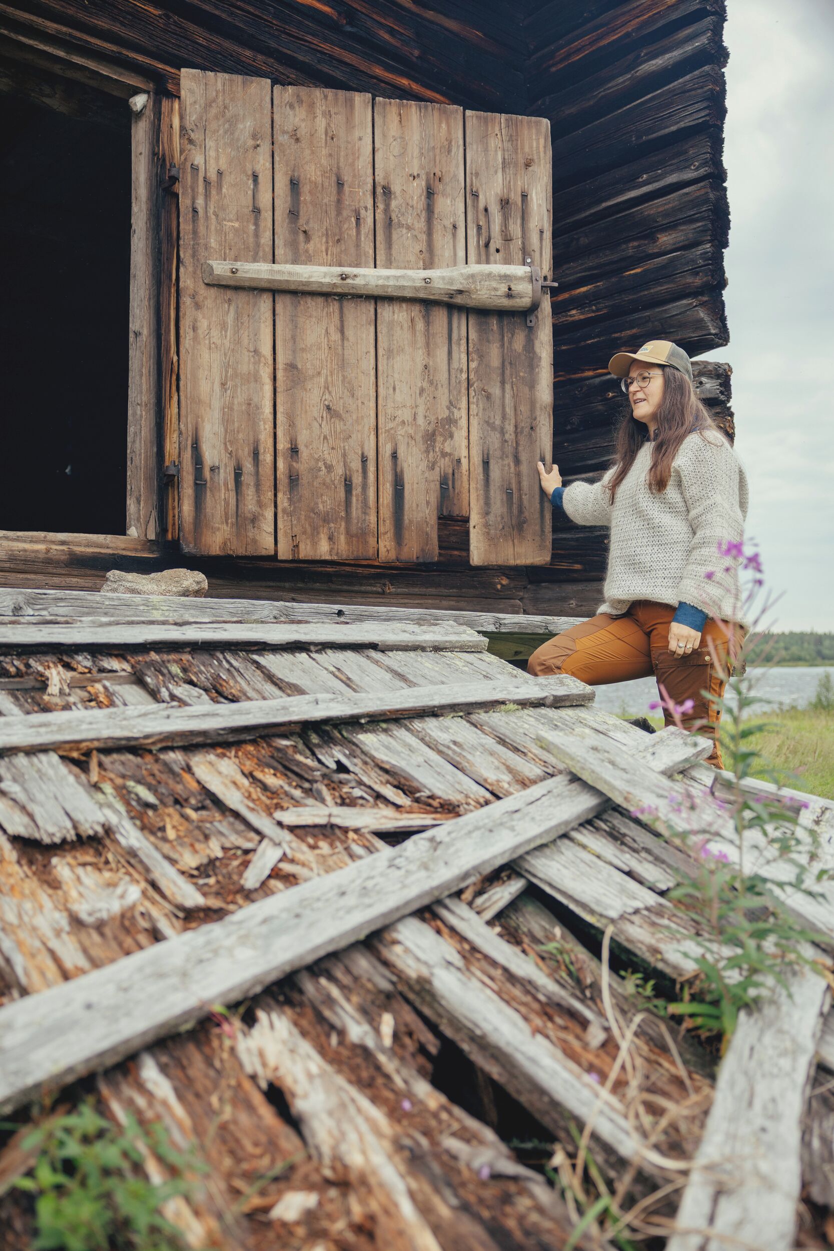 A rustic wooden cabin with a large door, a weathered wooden deck, and a person sitting on the deck.