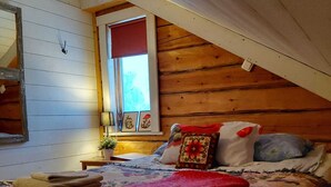 A cozy attic bedroom with a sloped ceiling, a bed with floral bedding, a red throw blanket, and a window with decorative items. - Lapland Nature & Health (Junosuando)