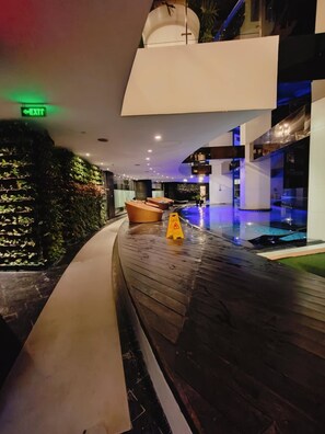 Un área de piscina interior moderna con suelo de madera curva, un letrero de salida verde y un letrero de precaución amarillo.