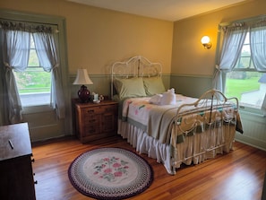 A bedroom with a wooden floor, a bed with a white metal frame, a nightstand with a lamp, and a window with curtains. - The Marr House on the River (Greene)