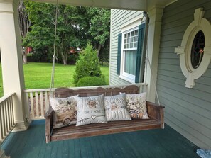 A porch with a wooden swing and cushions, a window with shutters, and a view of a green lawn and trees. - Romantic Historic Riverside Escape in the Village of Greene (Greene)