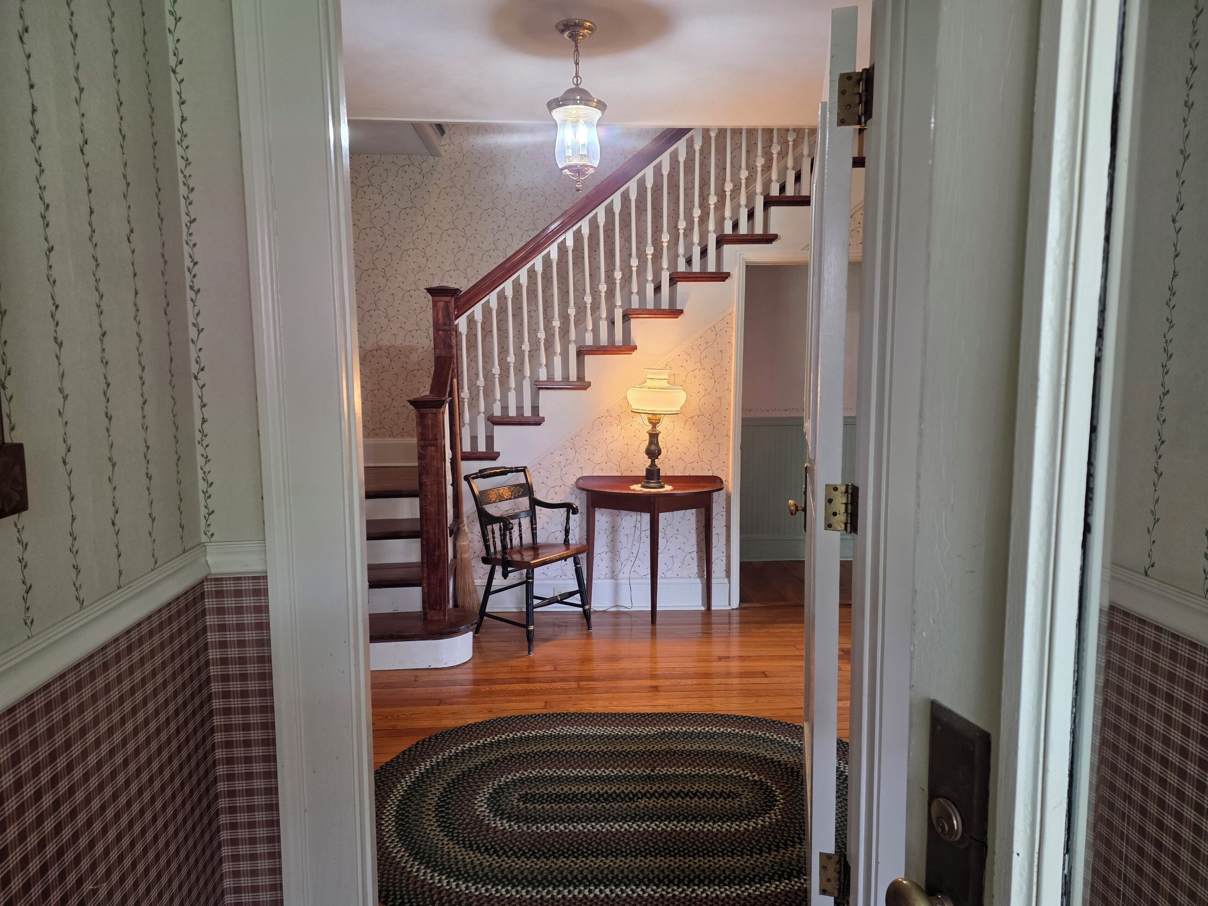 A staircase with a wooden handrail and white balusters, leading to a room with a patterned wallpaper and a table with a lamp.