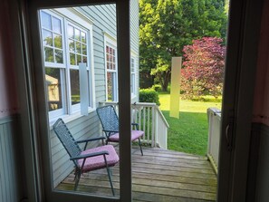 A wooden deck with two chairs and a table, overlooking a garden with a lawn and trees. - The Marr House on the River (Greene)