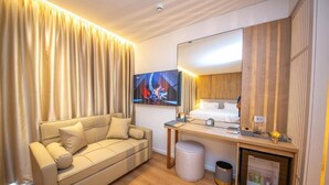 Habitación de hotel con sofá, cama, televisor, espejo y refrigerador pequeño.