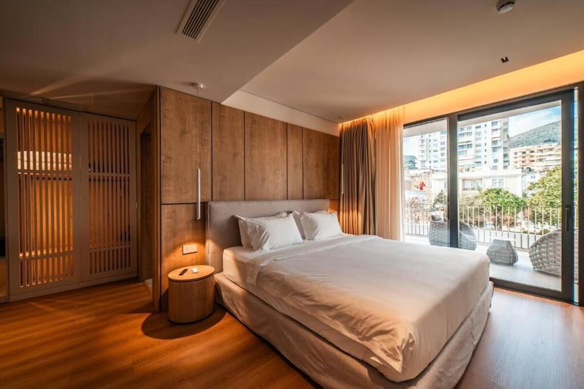 Un dormitorio moderno con una cama grande, una mesita de noche redonda, un armario de madera y un balcón con vistas a edificios.