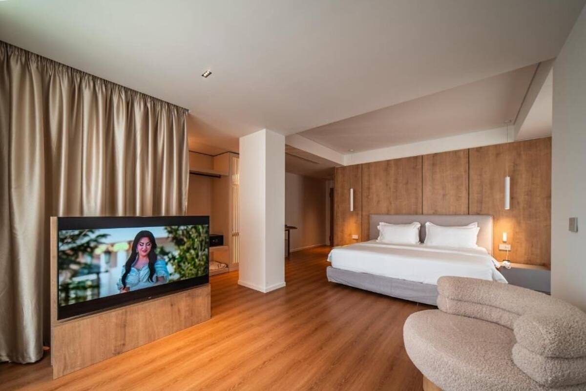 Habitación de hotel moderna con un televisor de pantalla plana grande, una cama con ropa blanca, cabecero de madera y una zona de estar con un sillón color beige.