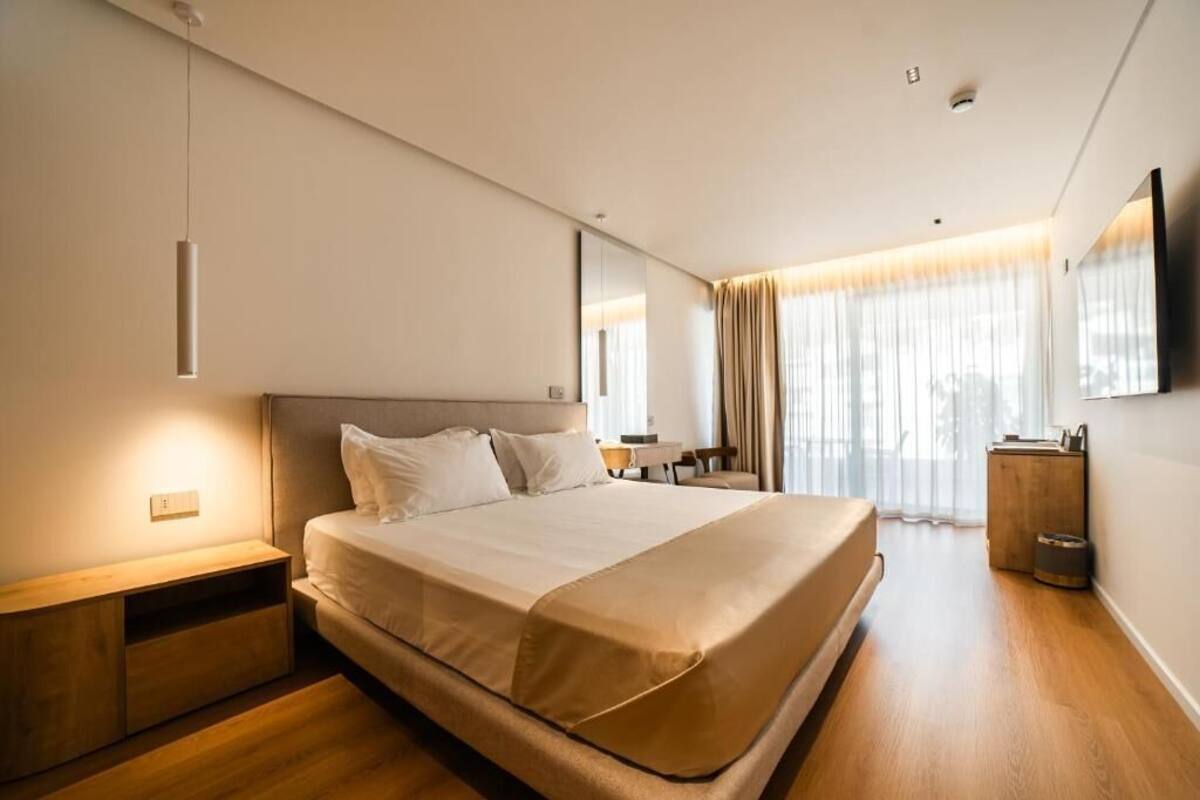 Una habitación de hotel moderna con una cama grande, mesitas de noche, un televisor de pantalla plana y un ventanal con cortinas.