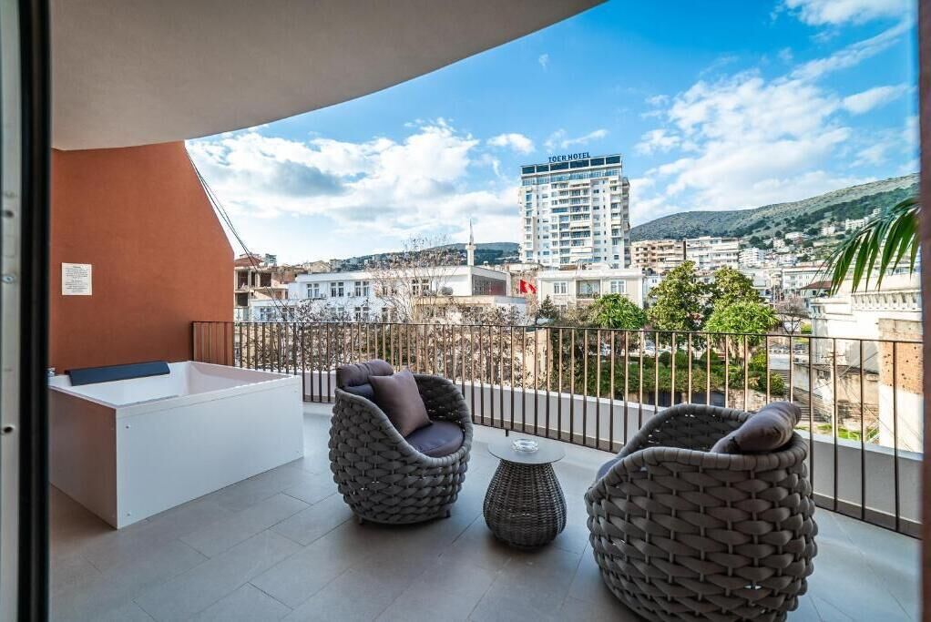 A balcony with a white bathtub, wicker furniture, and a view of a hotel and cityscape.