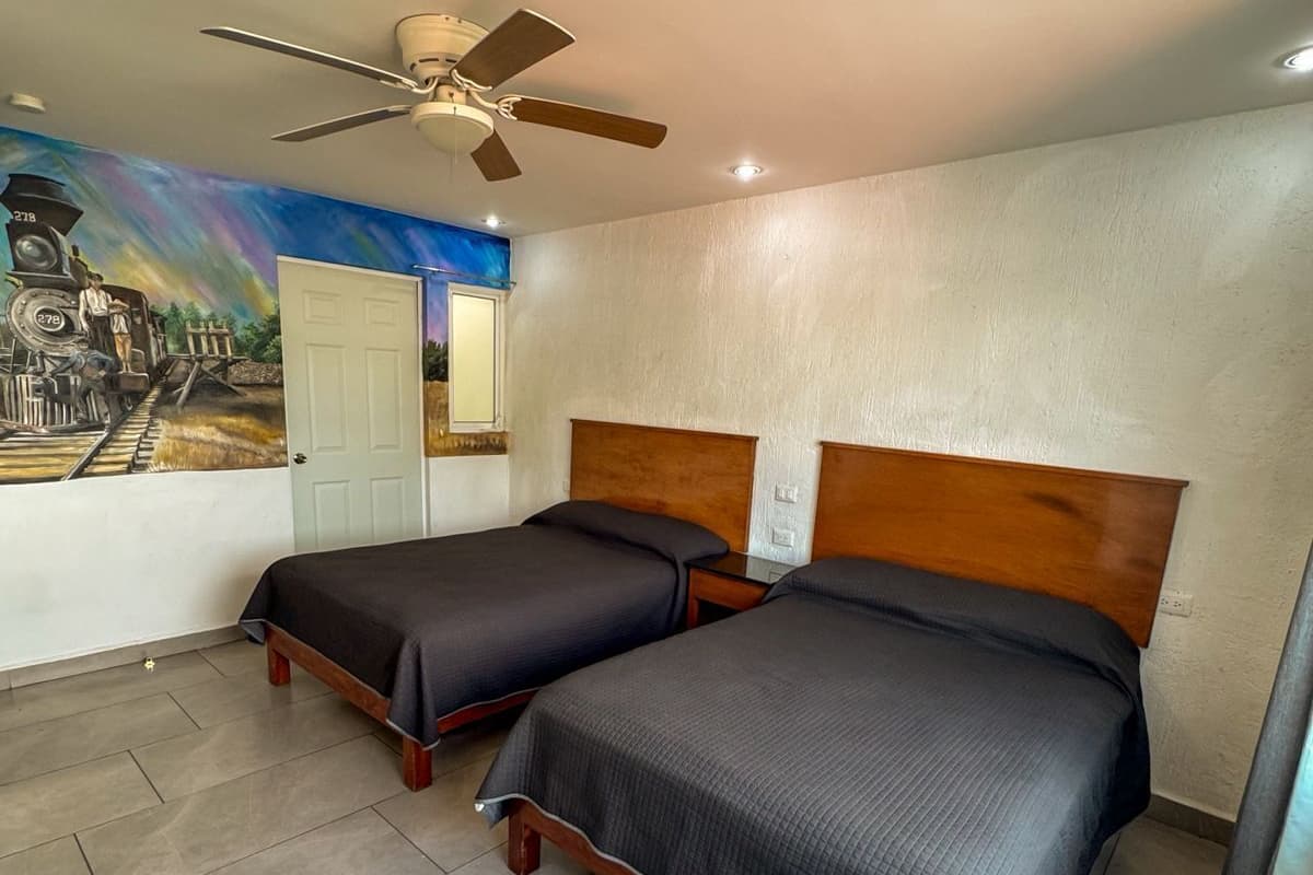 A hotel room with two beds, a ceiling fan, a mural on the wall, and a door.