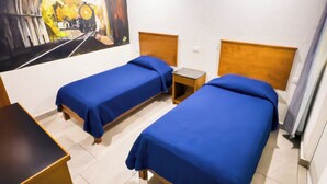 Une chambre d’hôtel avec deux lits simples, un ventilateur de plafond, un tableau représentant un tunnel de train et une petite table.