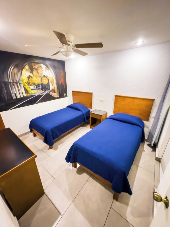 A hotel room with two single beds, a ceiling fan, a painting of a train tunnel, and a small table. - HOTEL CALIFORNIA (Soledad de Graciano Sánchez)