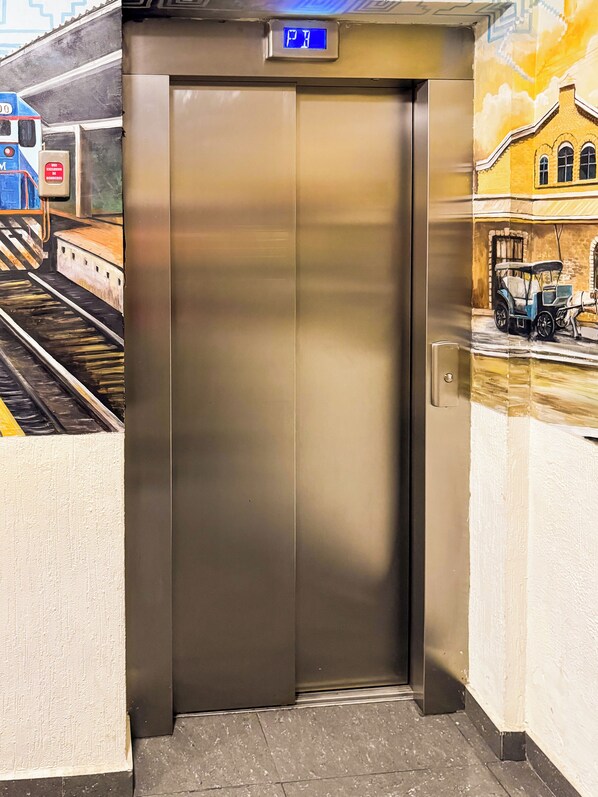Un ascensor con pantalla digital, piso de baldosas y un mural de un tren y un edificio en la pared.