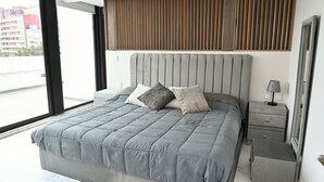 A modern bedroom with a large bed, a wooden panel headboard, a bedside table with a lamp, and a view of the city through large windows. - Luxury Penthouse in Polanco | Private Terraces + Prime Location (Ciudad de México)