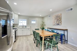 A modern kitchen with white cabinets, a stainless steel refrigerator, a wooden dining table with green chairs, and a blue chair at the counter.