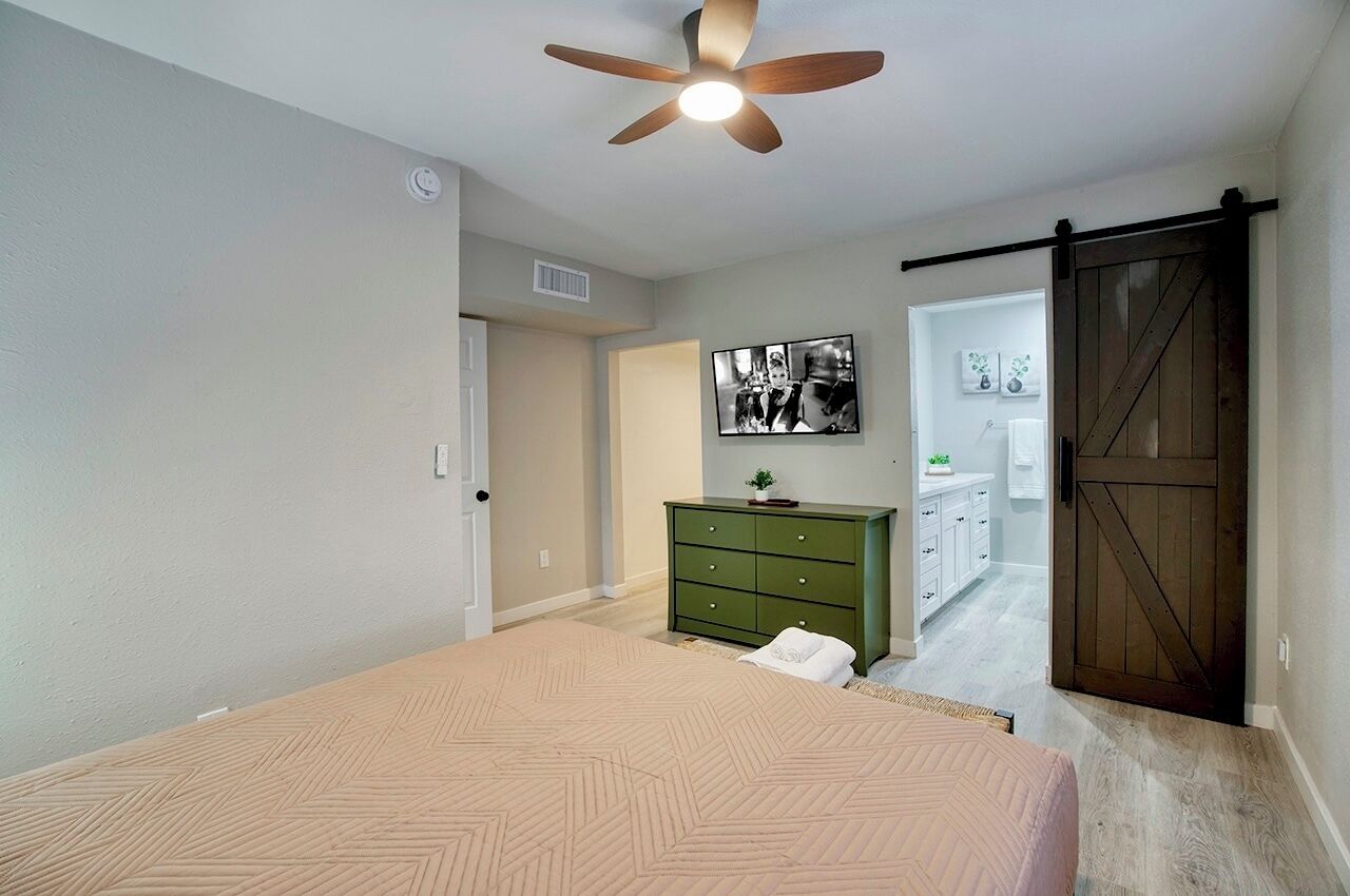 A bedroom with a bed, a dresser, a ceiling fan, a door, and a hallway leading to a bathroom.