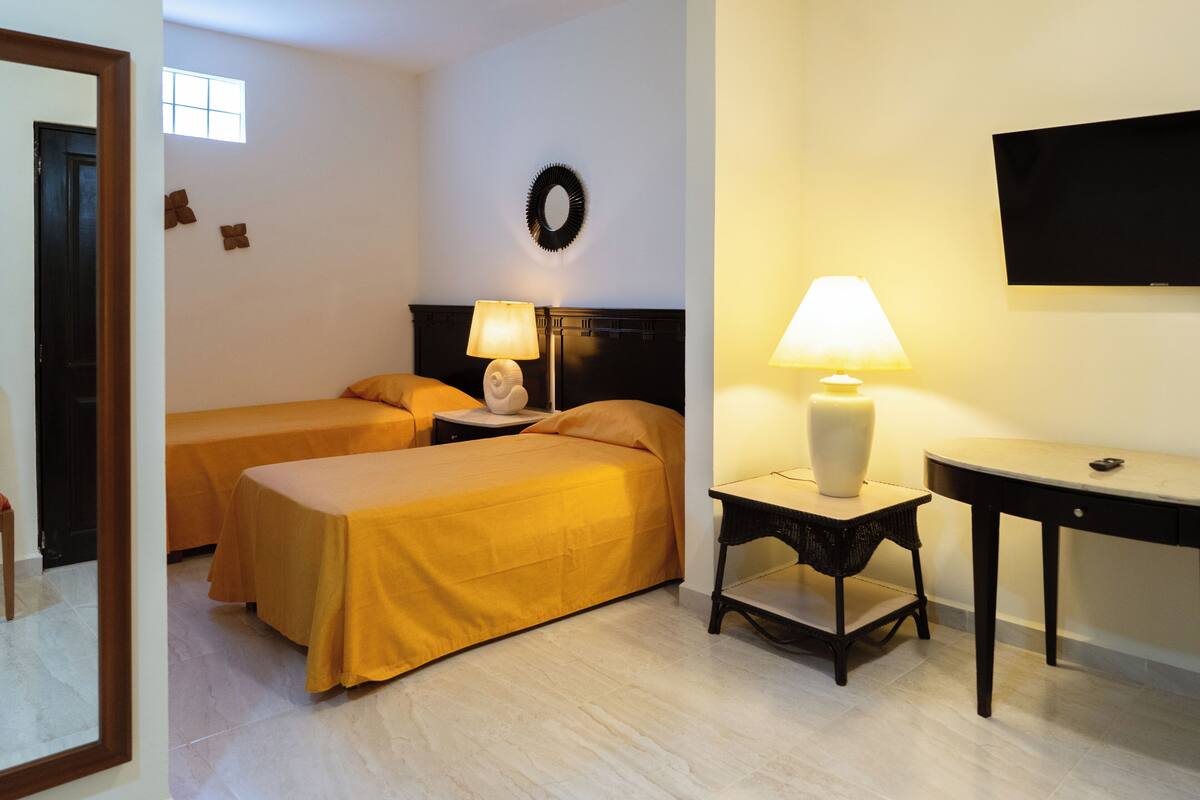 A hotel room with two beds, a nightstand with a lamp, a TV on the wall, and a small table.