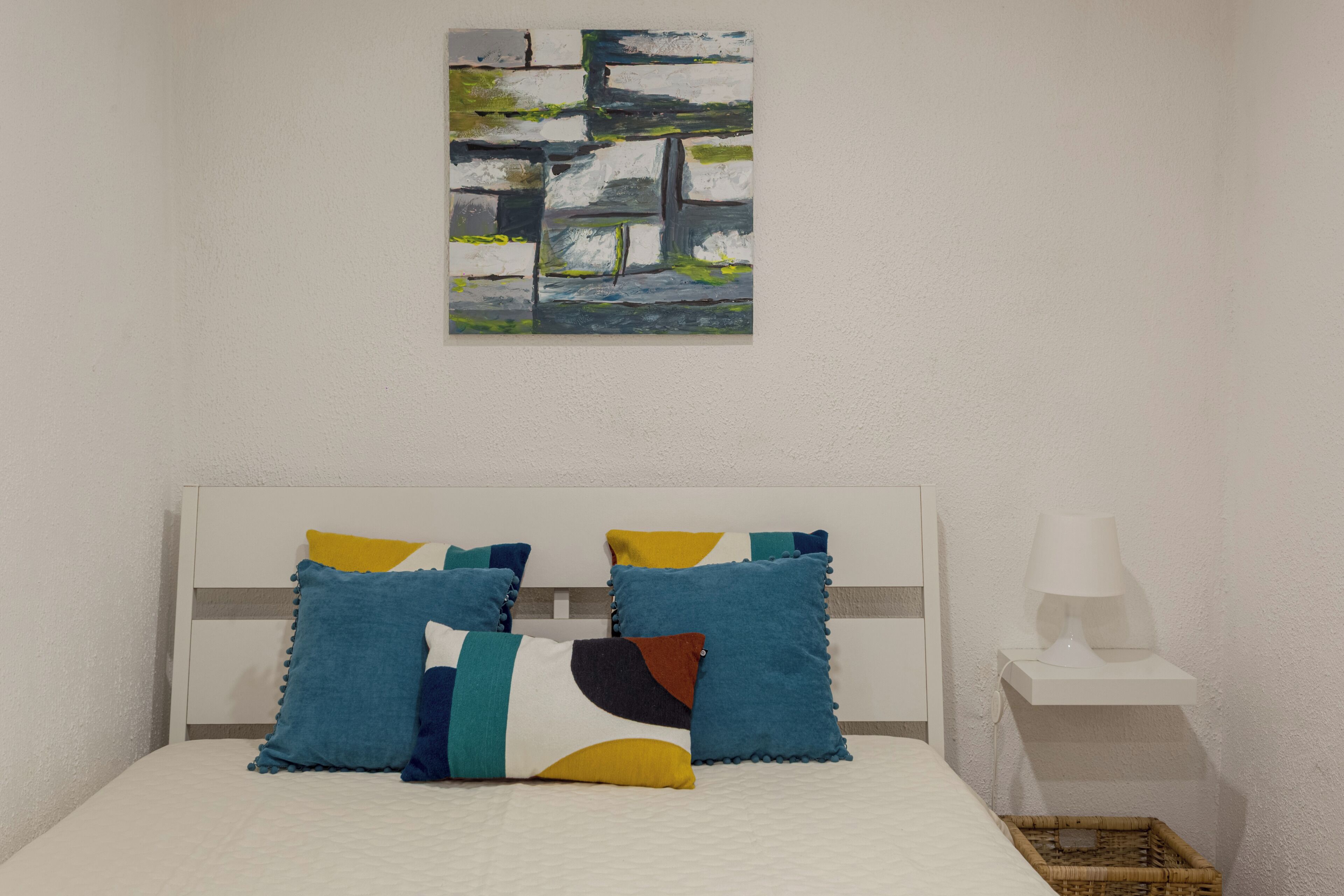 Ein Bett mit weißem Kopfteil, mehreren Kissen, einem Nachttisch mit Lampe und einem Wandbild im abstrakten Stil.