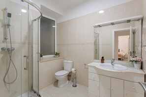 A bathroom with a glass-enclosed shower, a toilet, a sink with a mirror, and a window above the toilet. - Graça Secret Village – 2BR Retreat (Lisbon)