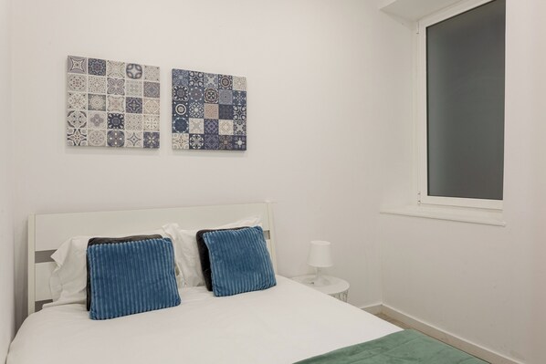 Une chambre à coucher comprenant un lit, des oreillers, une table de chevet avec une lampe, une fenêtre et deux œuvres d’art murales.