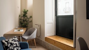 Een kleine kamer met een blauwe bank, een houten tafel, een grijze stoel, een kamerplant en een raam met uitzicht op gebouwen.