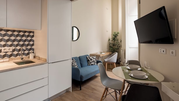 Een gezellig compact woongedeelte met een kitchenette, een eethoek met tafel en stoelen, een bank, een aan de muur bevestigde televisie en een potplant.
