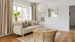 Una sala de estar con un sofá, un sillón de mimbre, una planta en maceta y un ventanal grande.